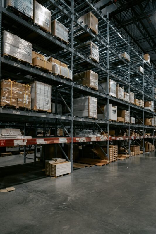 Tall warehouse racking aisle stacked with mixed wrapped pallets and boxes